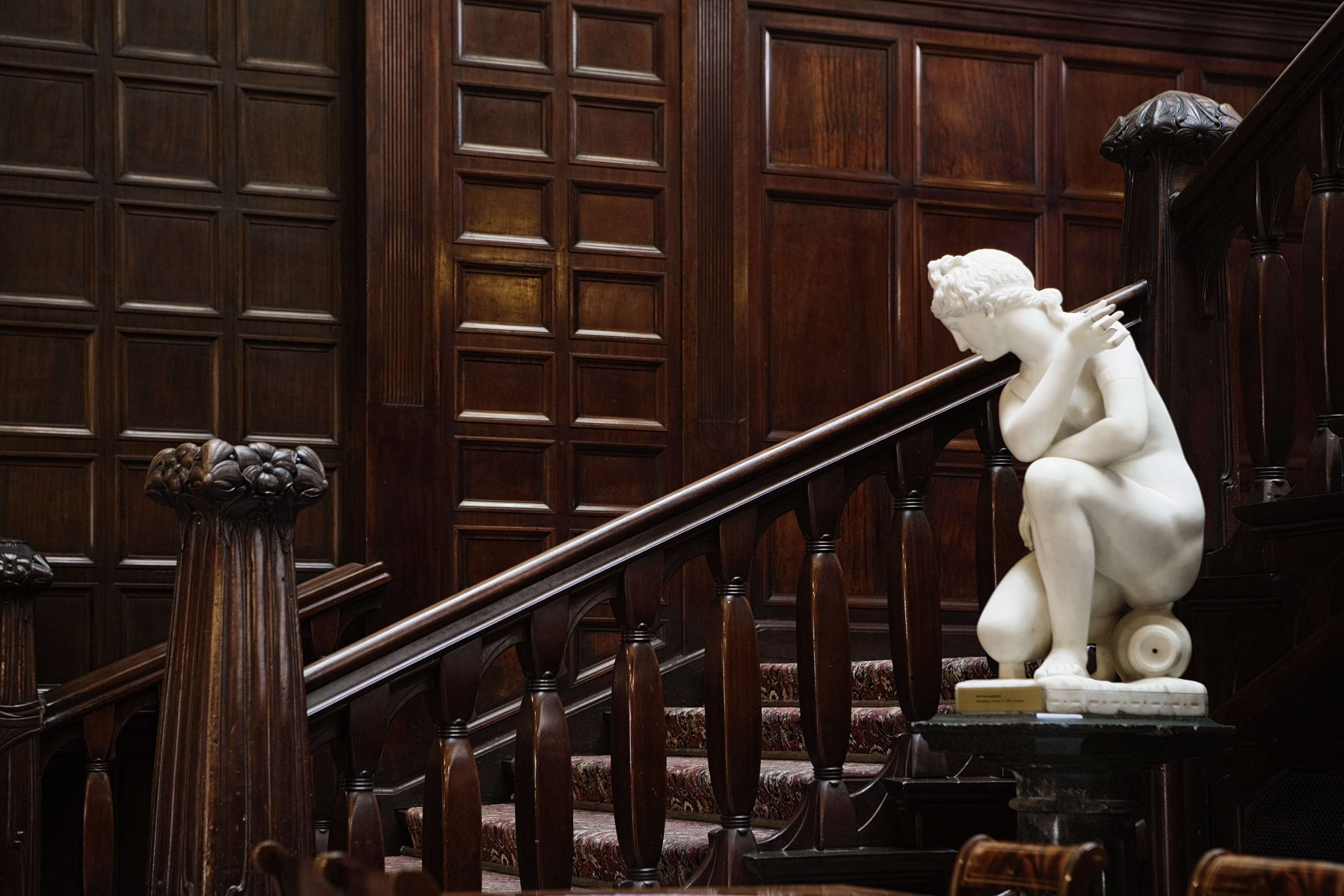 Widok na drewniane schody z ozdobna poręcza i alabastrowa rzeźba postaci kobiecej na postumencie.
