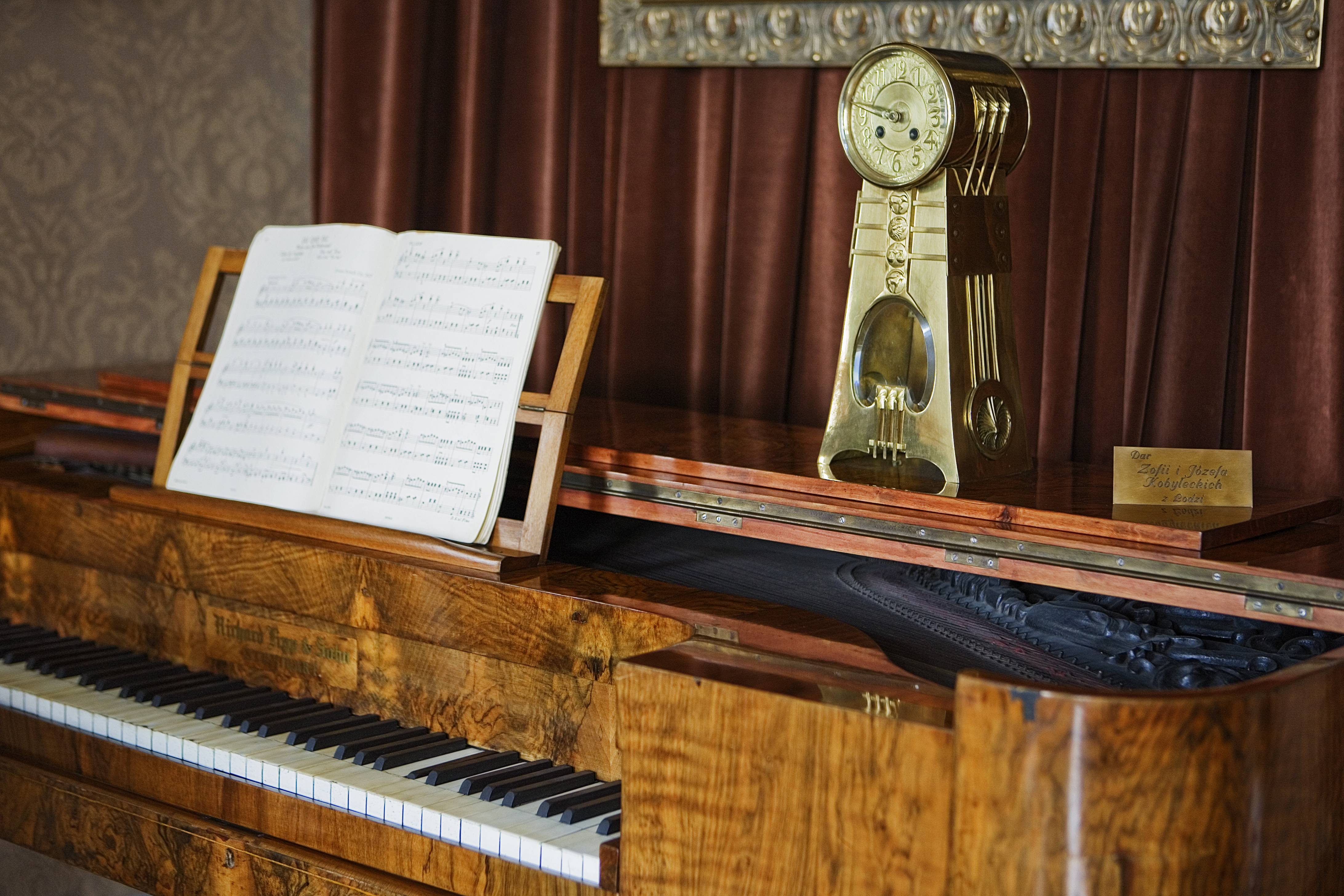 Zbliżenie na fortepian salonowy z jasnego drewna, z uchylona klapa i stojący na nim zabytkowy zegar mechaniczny.