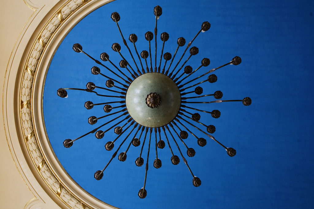 Widok sufitu sali lustrzanej ze sztukateria, intensywnym niebieskim malowaniem owalnej części centralnej i wieloramiennym żyrandolem elektrycznym.