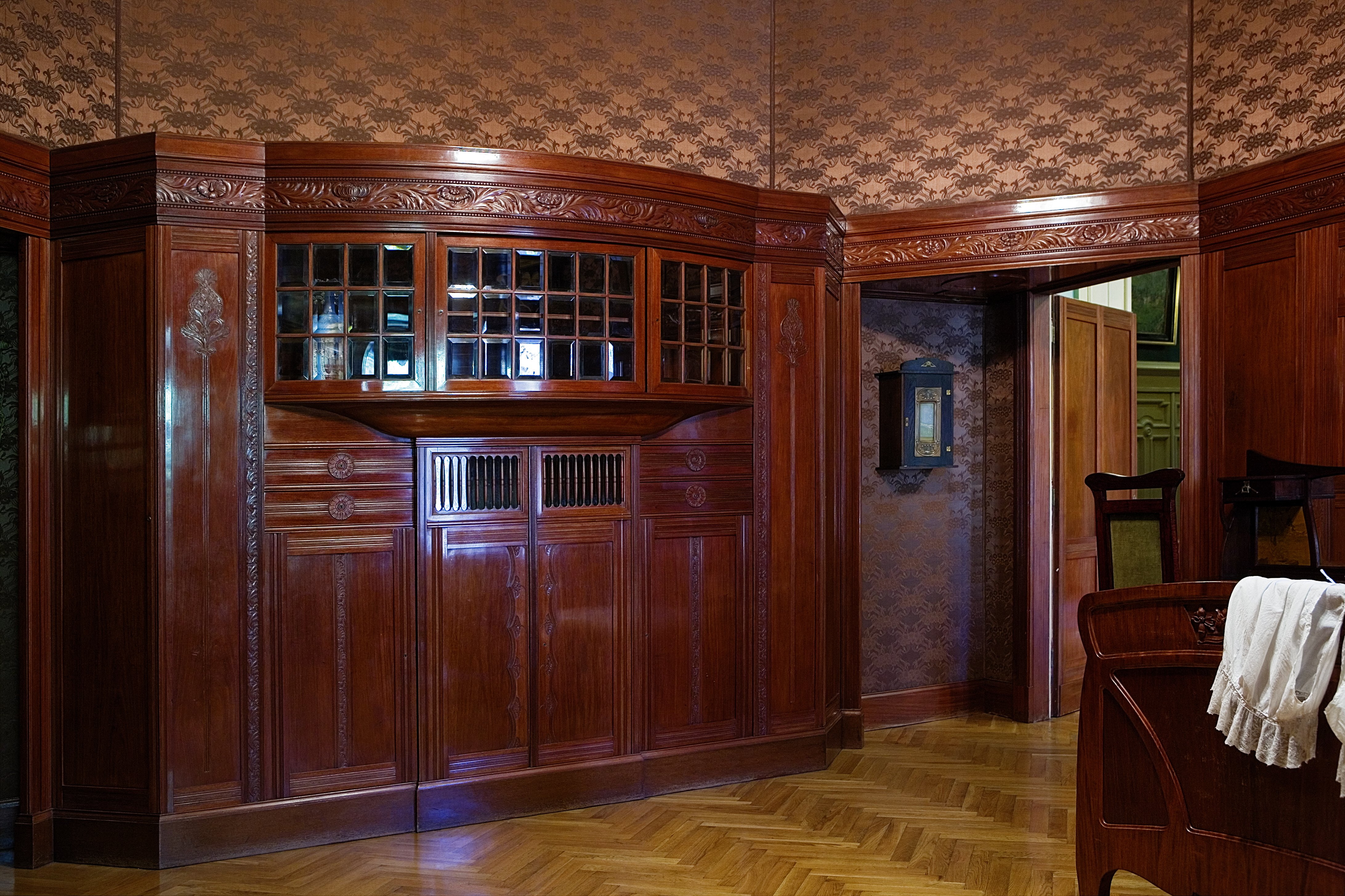 Widok na wyposażenie sypialni pani domu - drewniane, rzeźbione szafy, wbudowane w boazerię w kolorze mahoniowego drewna, przeszklone niewielkimi kryształowymi szybkami.
