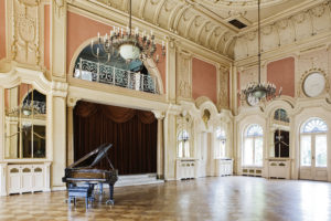 Widok sali balowej pałacu, bogato dekorowanej płaskorzeźbami i licznymi lustrami, z fortepianem na pierwszym planie.