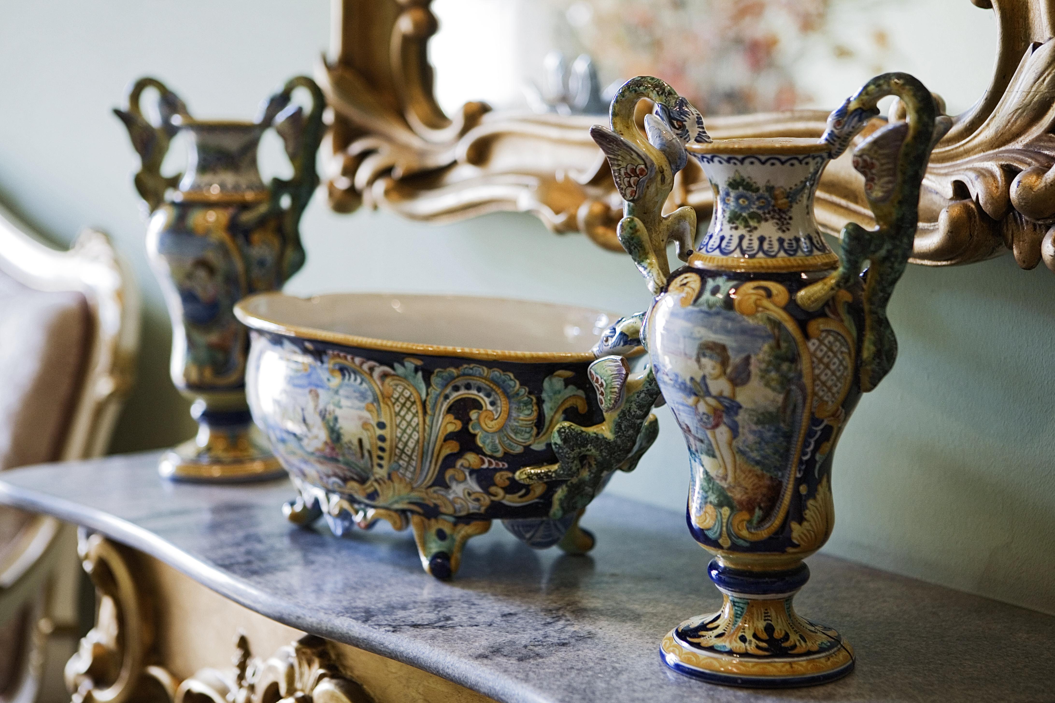 Widok bogato zdobionych wazonów ceramicznych z uchwytami w kształcie smoków w dominujących w kolorach złotym i niebieskim, na kamiennej półce pod lustrem w rzeźbionej ramie.