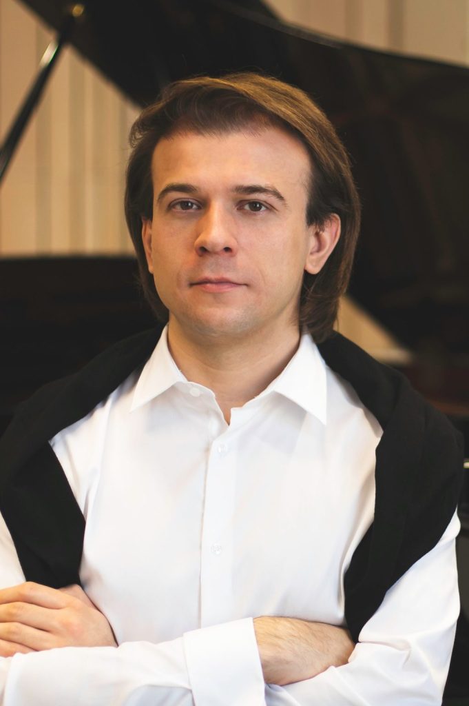 Zdjęcie mężczyzny w białej koszuli z narzuconym na ramiona czarnym swetrem. Mężczyzna stoi z założonymi rękami w tle widać fortepian.