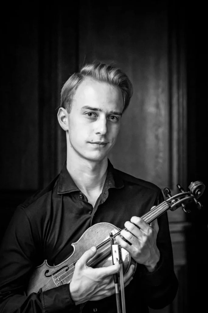 Czarno-białe zdjęcie uśmiechniętego mężczyzny. W rękach na wysokości klatki piersiowej trzyma skrzypce i smyczek.