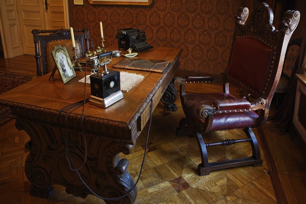 Zbliżenie na rzeźbione biurko i wyściełany fotel fabrykanta Izraela Poznańskiego, z urządzeniami biurowymi z końca XIX wieku: bakelitowym telfonem, maszyną do pisania oraz przyborami do pisania piórem atramentowym.