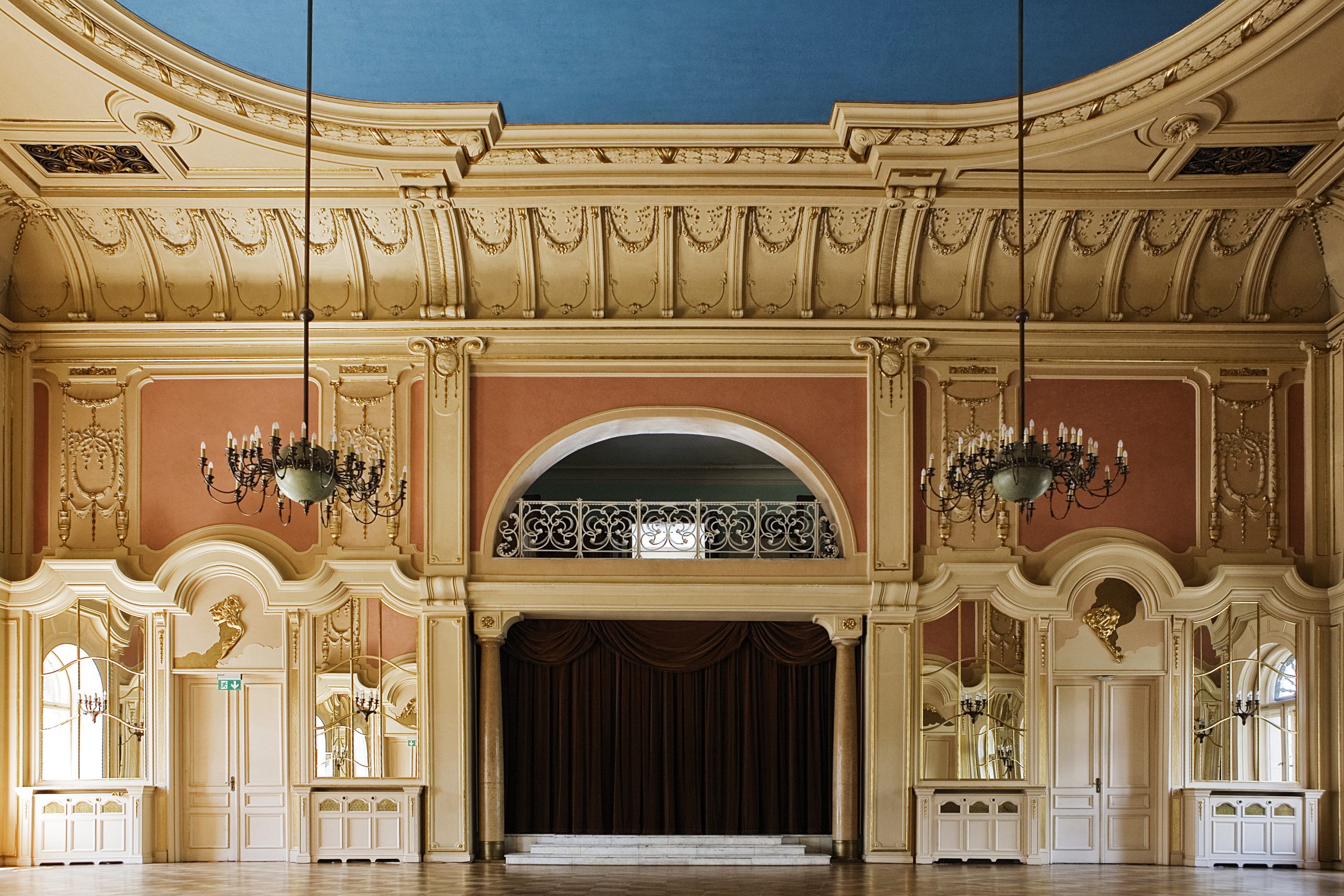 Widok jednej ze ścian bogato zdobionej sali lustrzanej z wejściami do przylegających pomieszczeń oraz wnęki o charakterze sceny, przesłoniętej ciężka, ciemna kurtyna.